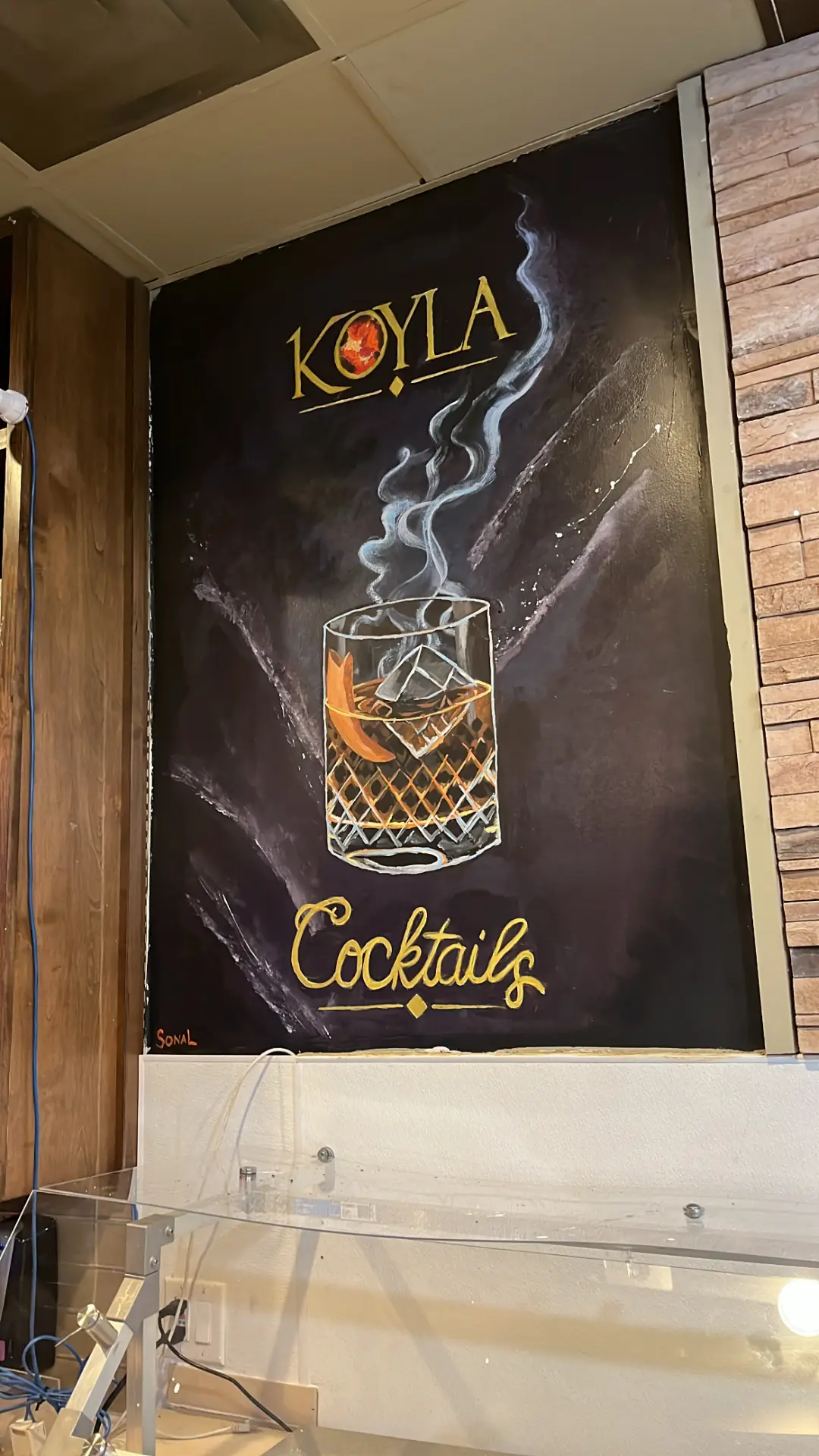 Hand-painted Koyla Cocktails mural with gold lettering and a smoking whiskey glass on a black bar wall